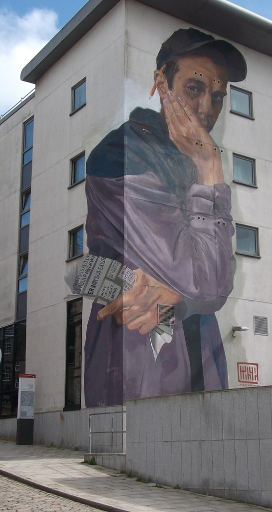 A man with a cap, pencil behind his ear and a flier in his hand, wearing a grey jacket and looking pensive