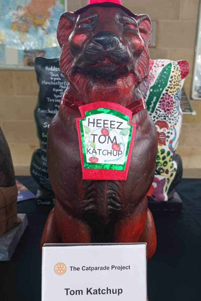 A red tinted cat statue with sticker on stomach stating Tom Katchup