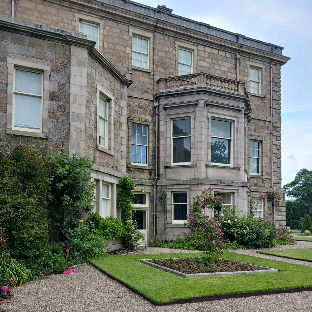 A mansion house with dormer windows and balustrades, and a square of grass with roses in front.