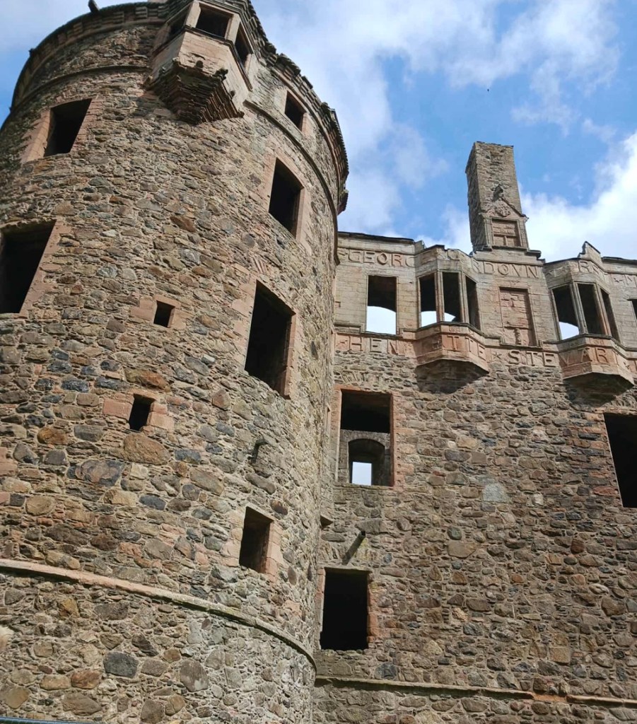 A ruined castle with a rounded section of stone wall and a straight wall with rectangular windows