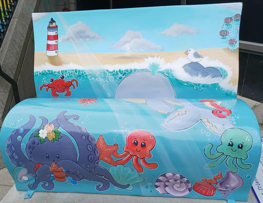 A blue bench with paintings of sea creatures including an octopus, sea snail, jellyfish, crab and dolphin. Beyond the sea there is sand and a lighthouse