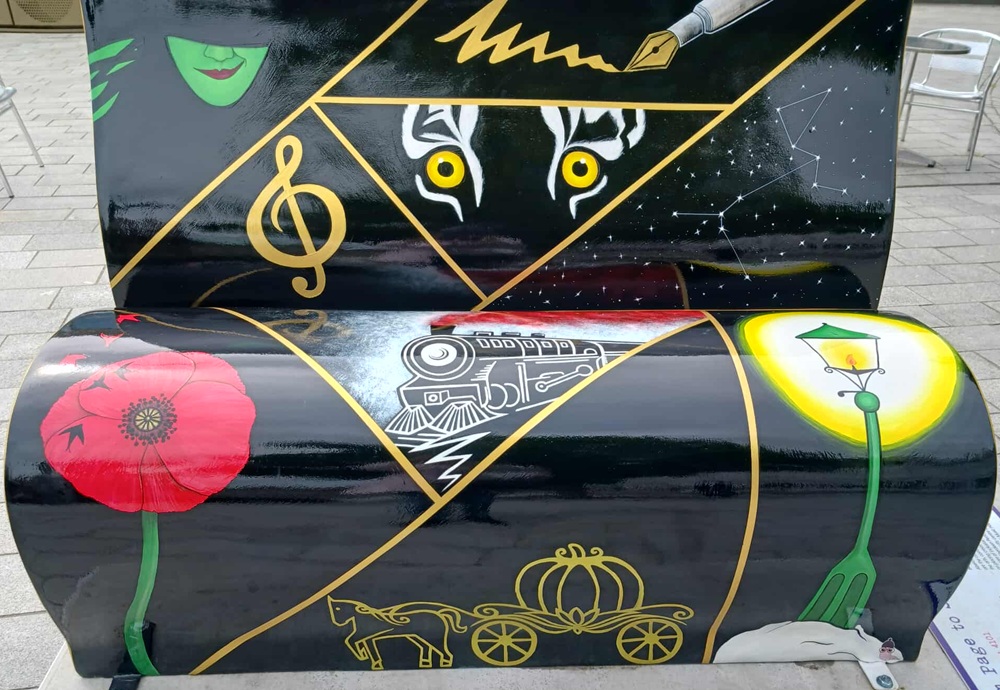 A black bench featuring images of a witch, a fountain pen, a musical note, the eyes of a cat, a steam train, a red poppy, a pumpkin and a streetlight