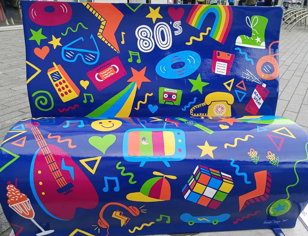 A bright blue painted bench with images from the 1980s including colourful cassette tape, record, glasses, giant mobile phone, landline telephone with dial, TV with aerial and guitar
