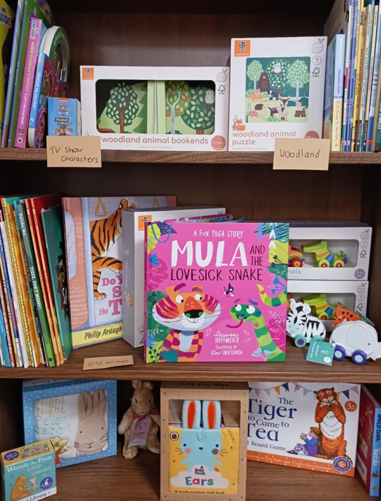 Colourful children's books on wooden shelves, the most prominent one is Mula and the Lovesick Snake