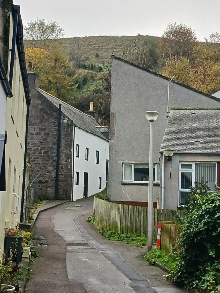 A narrow alley between stone houses with a hill and trees above