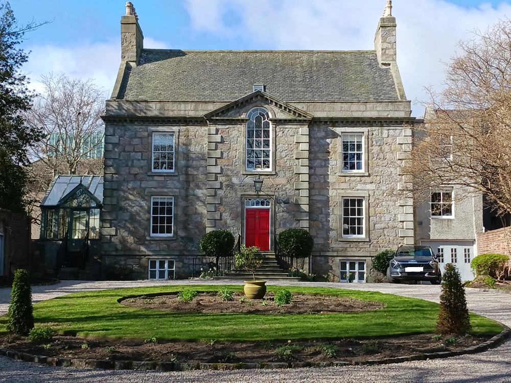 A stately house with two chimneys on either side, four grid windows and a larger central grid window beneath an ornate triangle structure and with a red door below. A circular drive surrounds a grass feature.