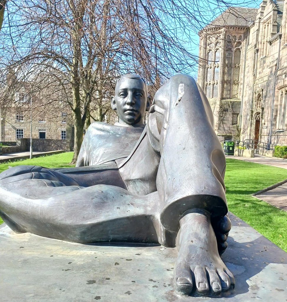 A bronze sculpture of a man sitting with one leg crossed and one with raised knee. There is a tree behind and old buildings behind and to the right.