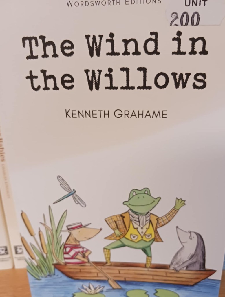 A book titled The Wind in the Willows with a picture of a frog in a coat in a boat with a rat and a mole.